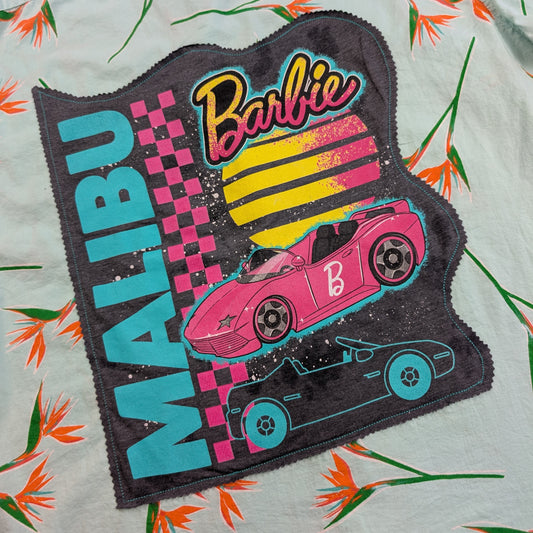 A close-up of a Barbie-themed fabric patch laid flat on a light-colored floral surface. The patch is dark gray with a scalloped edge and features the word 'MALIBU' in large turquoise letters, a pink and yellow striped sun, a pink race car, a black race car, and the Barbie logo in pink and yellow.