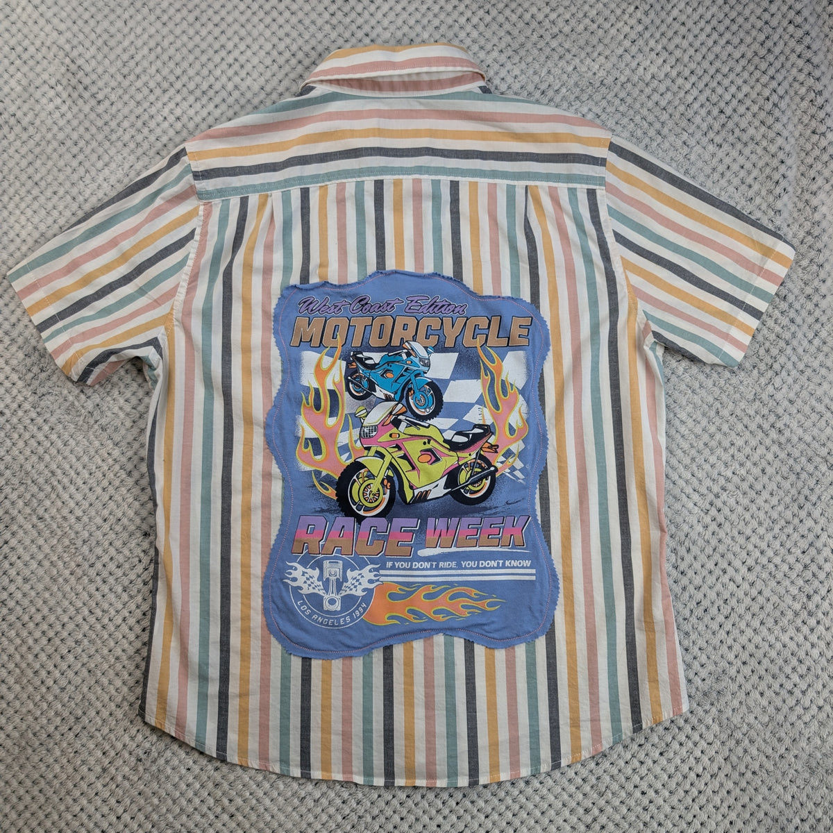 A short-sleeve button-up shirt with vertical stripes in beige, orange, green, and gray, featuring a large blue graphic print on the back with illustrations of motorcycles, flames, and the text 'MOTORCYCLE RACE WEEK'.