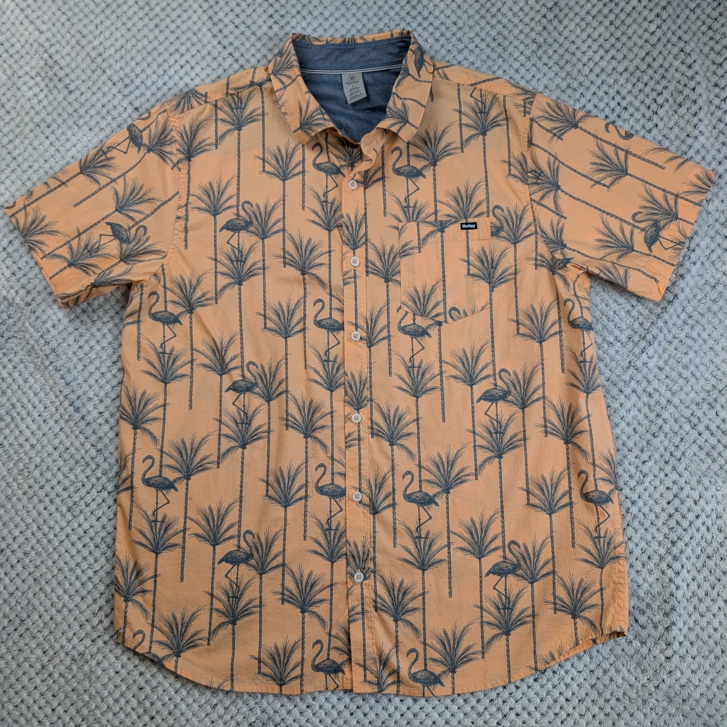 A short-sleeve button-up shirt with a tan and navy blue tropical flamingo and palm tree pattern, laid flat on a gray textured background.