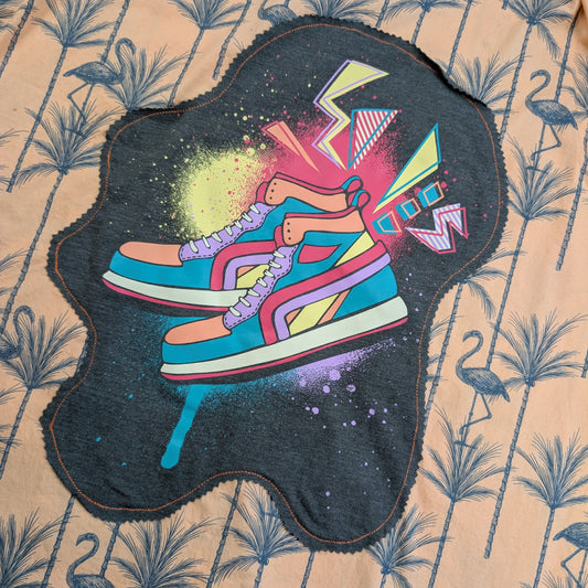 A dark charcoal gray fabric patch featuring a colorful graphic of two sneakers with abstract paint splatters and geometric shapes, laid flat on a beige tropical flamingo-patterned fabric background.