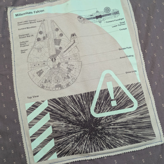 A close-up of a printed textile or patch featuring a Millennium Falcon diagram, sci-fi line art, and a large warning-style exclamation mark on a patterned gray fabric background.