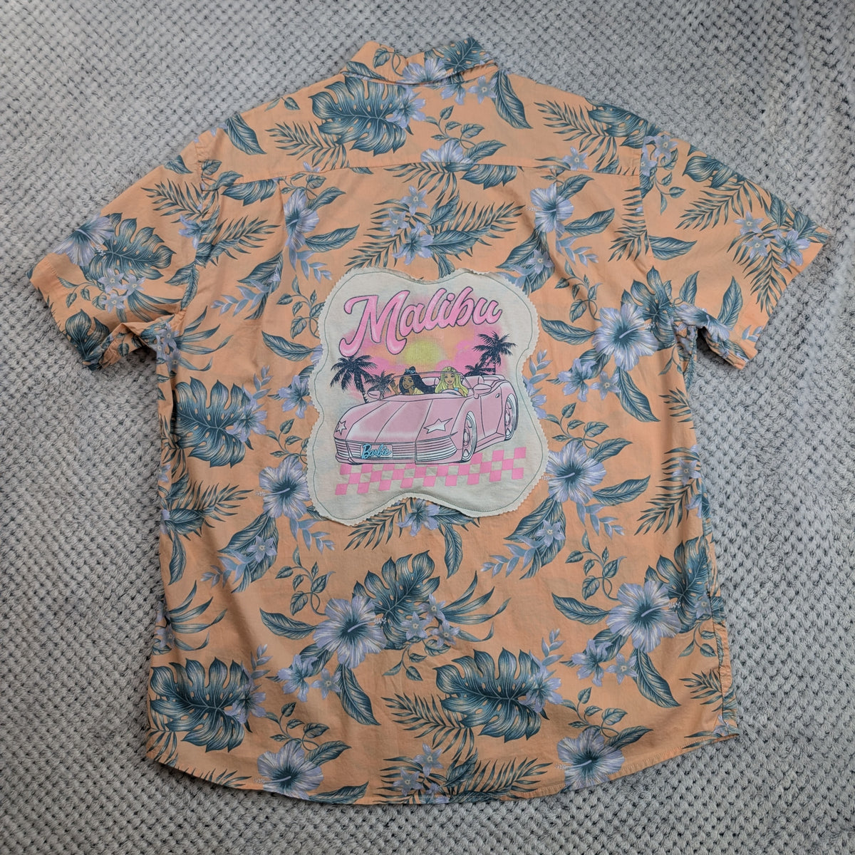 A peach-colored short-sleeve Hawaiian shirt with a blue and white floral print, featuring a large 'Malibu' graphic patch on the back with a pink car and palm trees, laid flat on a gray textured surface.