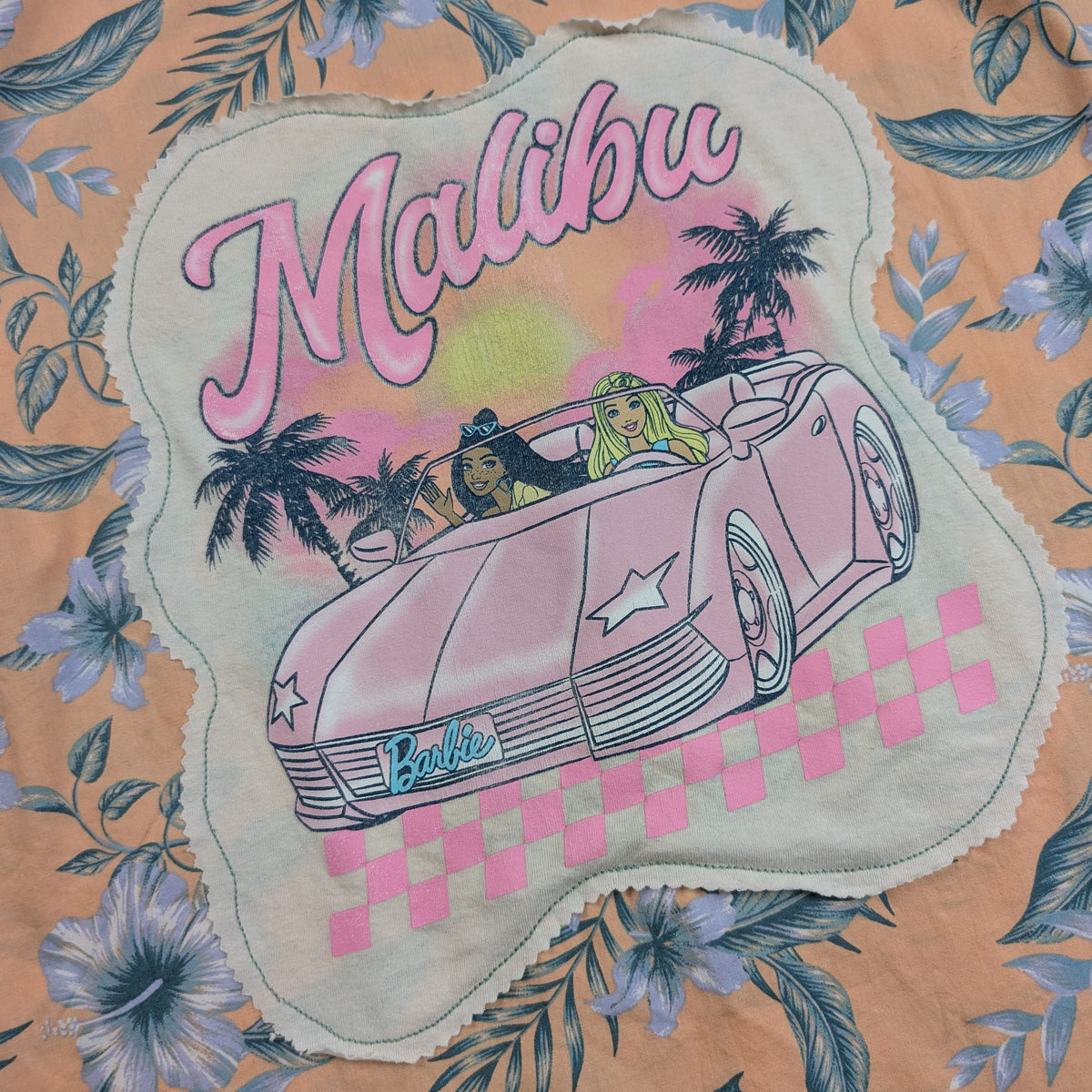 A close-up of a floral fabric featuring a scalloped applique patch with a vintage-style illustration of a pink convertible car, palm trees, and the words 'Malibu' and 'Barbie'.