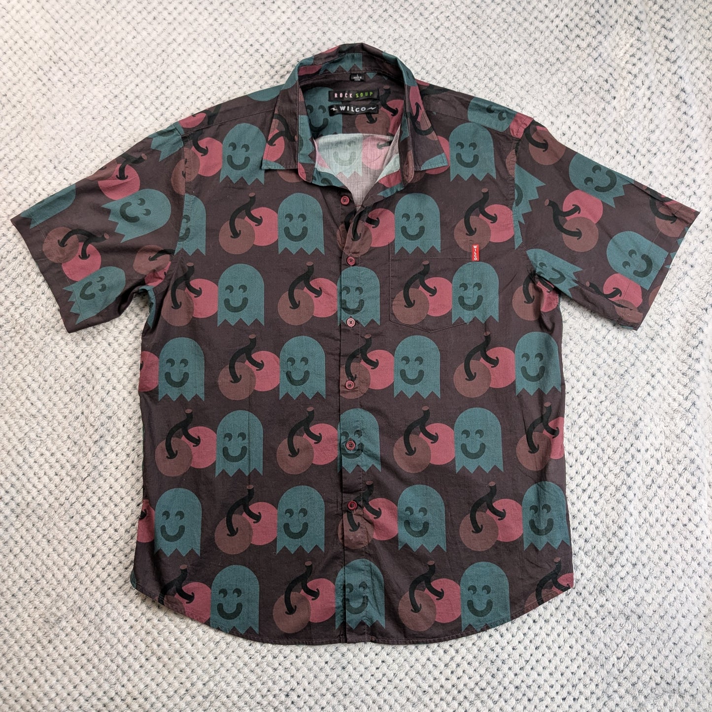A short-sleeve button-up shirt featuring a repeating pattern of smiling ghosts in teal and pink on a dark brown background, laid flat on a textured white surface.