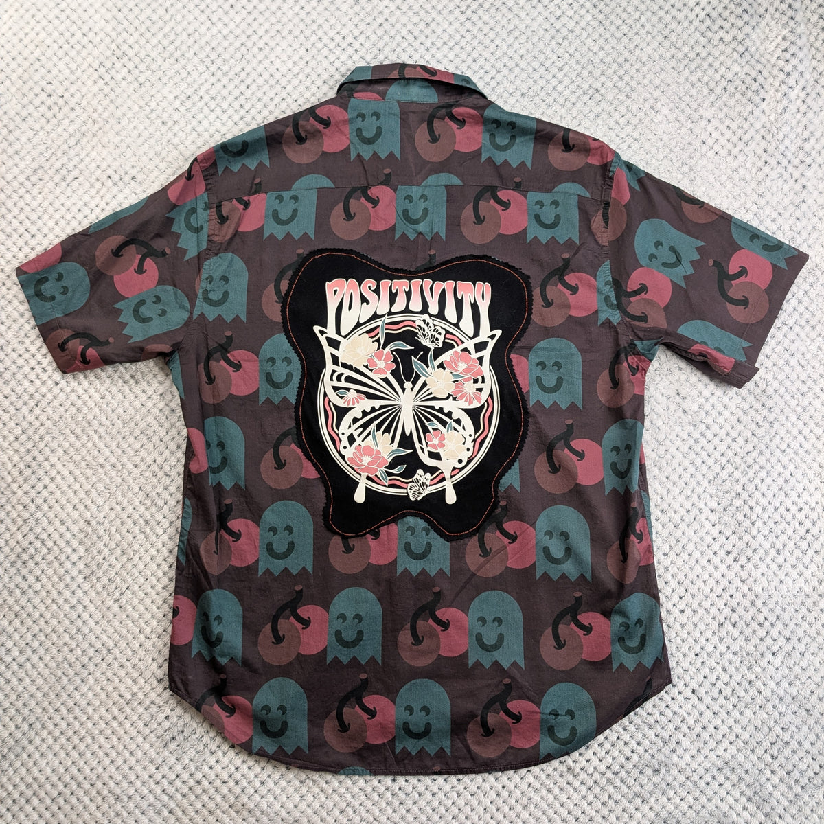 A short-sleeve button-up shirt laid flat on a textured light gray surface, showing the back side. The shirt has an allover print of pink and teal ghosts on a dark brown background, with a large black graphic patch in the center featuring a butterfly, flowers, and the word 'Positivity'.