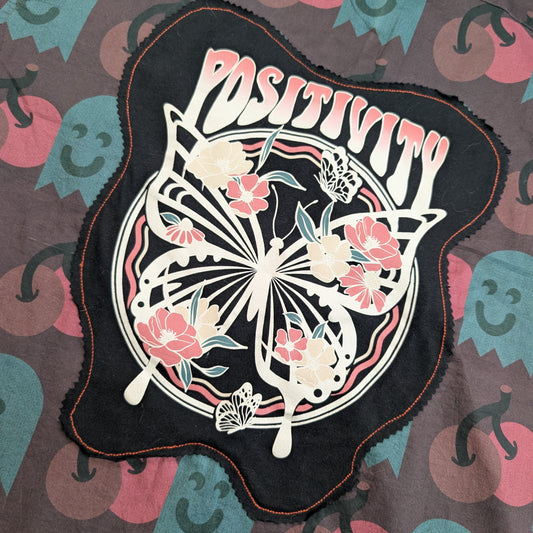 A black fabric patch with a scalloped edge featuring a butterfly and floral design with the word 'POSITIVITY' in pink and white, laid flat on a patterned fabric background.