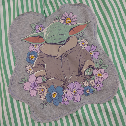 A gray fabric patch featuring a sleeping green cartoon character surrounded by purple, blue, and white flowers, placed on a green and white striped background.