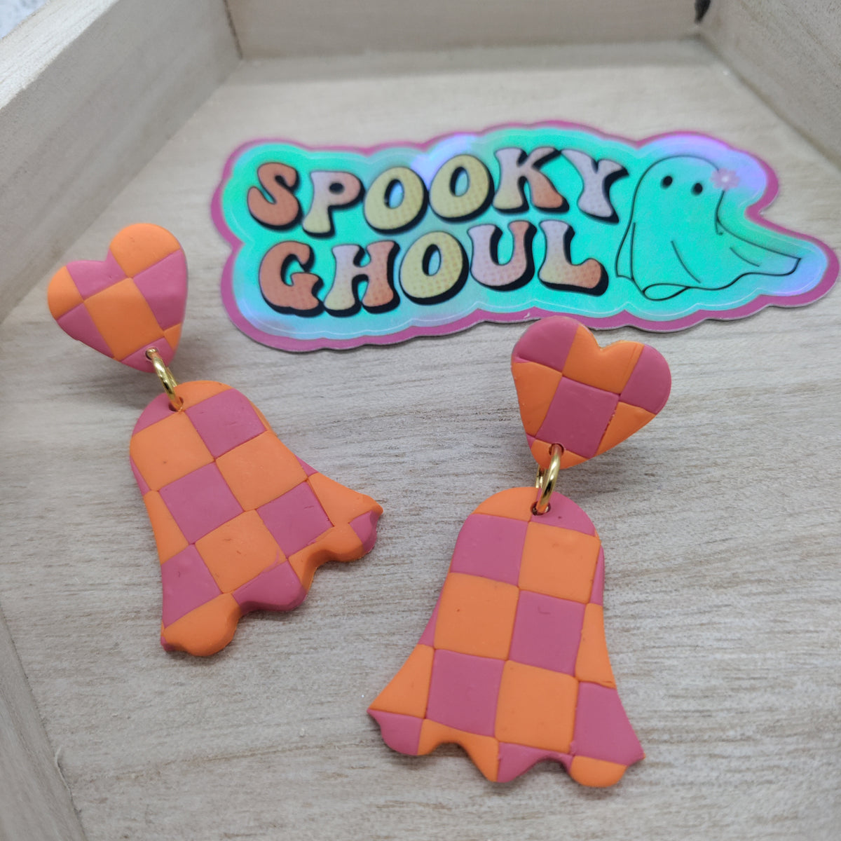 A pair of handmade ghost-shaped dangle earrings with a pink and orange checkerboard pattern, displayed on a light wood surface next to a 'Spooky Ghoulish' sticker.