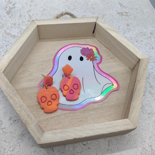 A pair of skull-shaped dangling earrings displayed in a hexagonal wooden tray with a ghost sticker on a light stone surface.