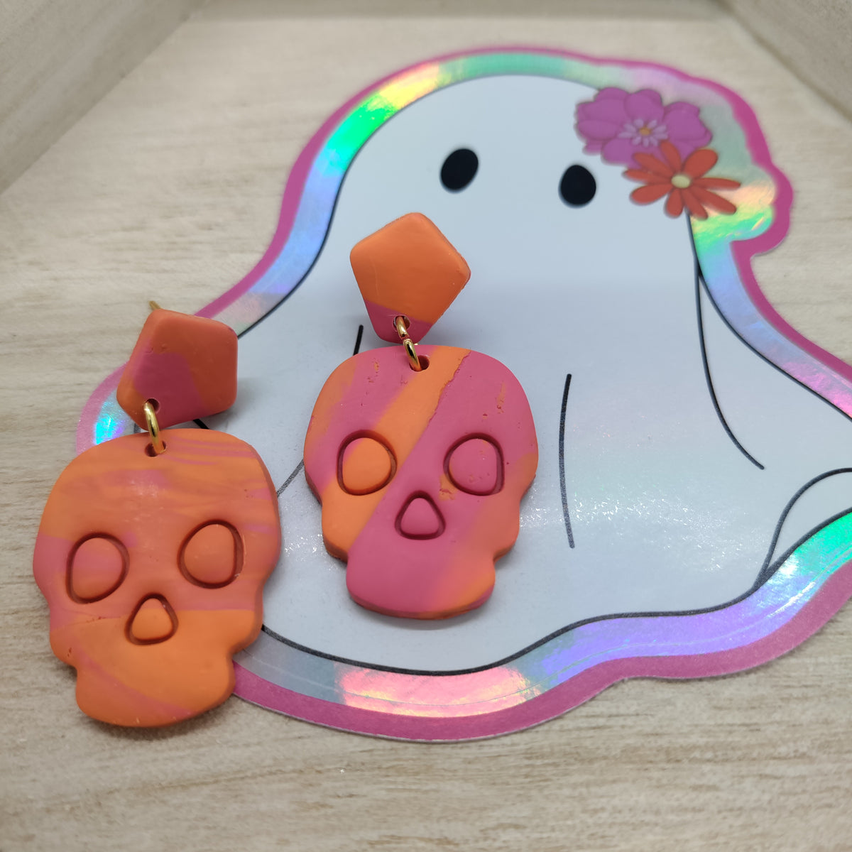 A pair of handmade drop earrings shaped like stylized skulls, featuring a color-blocked orange and pink design with gold-tone rings, displayed on a white ghost-shaped sticker over a light wood surface.