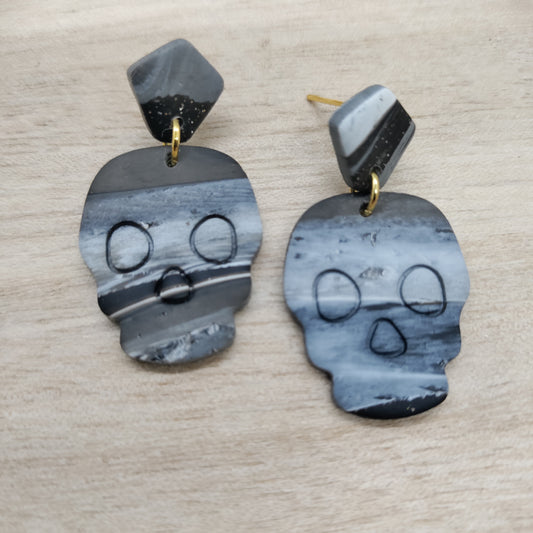 A pair of skull-shaped drop earrings made from polished black and white marbled stone, featuring gold post backings, displayed on a light wood background.