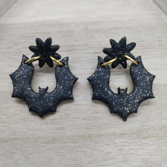 A pair of black bat-shaped hoop earrings with gold-tone ear loops and a small gold star detail on top, displayed on a light wood background.