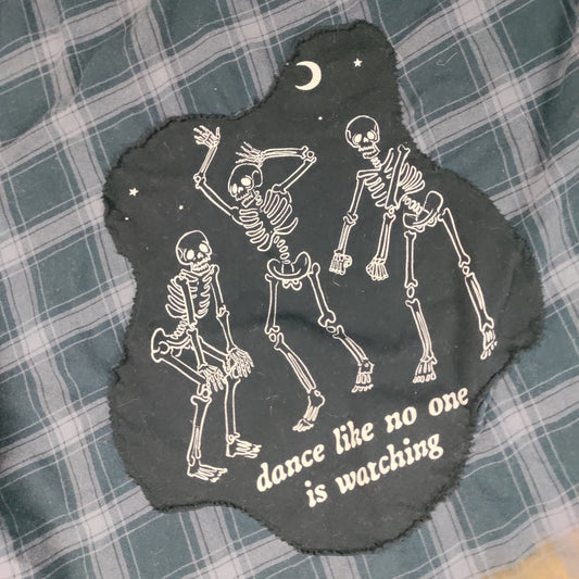 Black patch with skeletons and 'dance like no one is watching' text on a plaid fabric background
