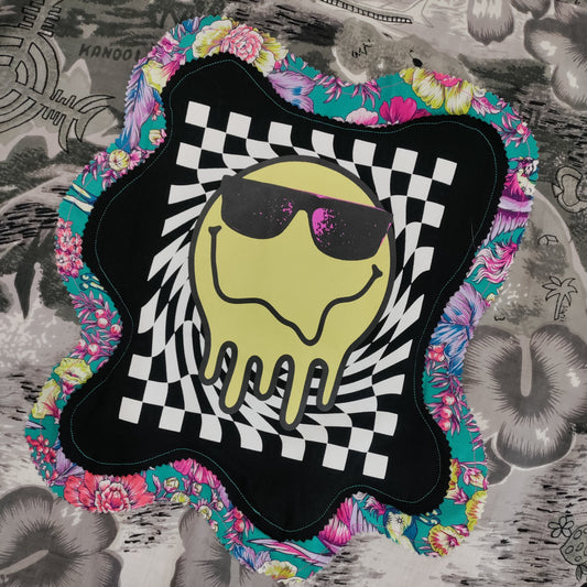 Decorative patch with a smiley face wearing sunglasses on a checkered background, surrounded by colorful floral patterns on a textured surface.