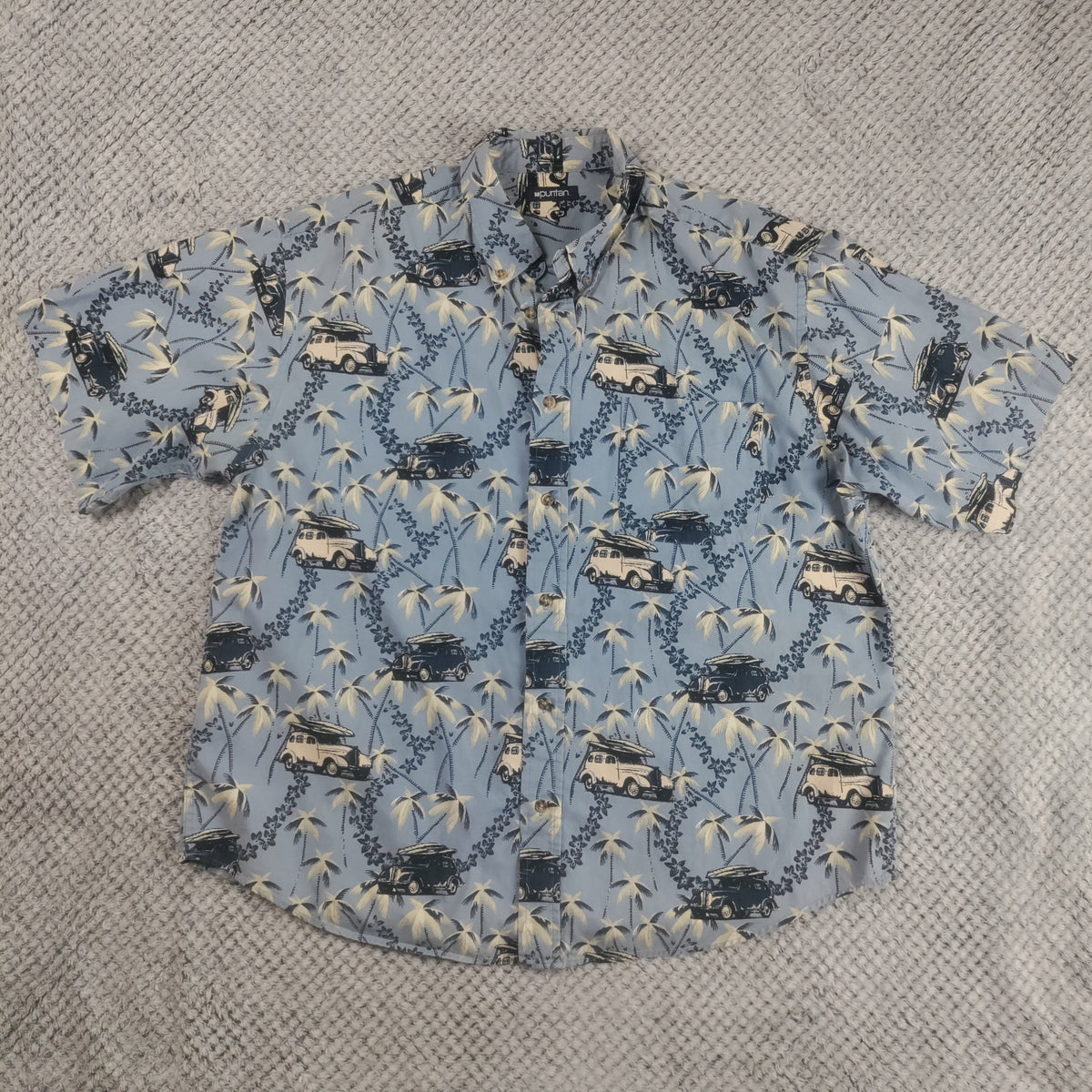 A short-sleeve button-up shirt with a light blue background and a repeating pattern of palm trees, and vehicles with surfboards, laid flat on a gray textured blanket.