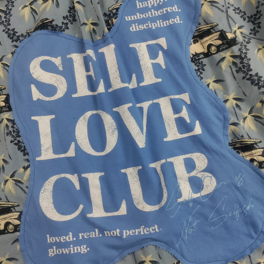 A blue fabric patch with a scalloped edge featuring the text 'SELF LOVE CLUB' in distressed white letters, laid flat on a gray tropical palm tree print fabric.