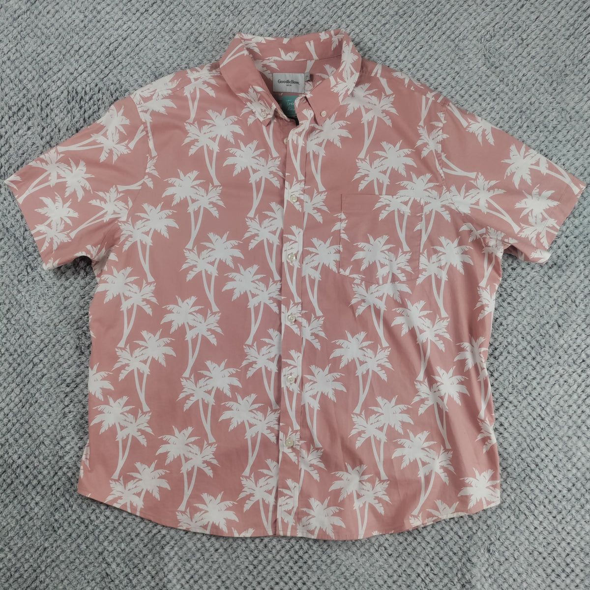 A short-sleeve button-up shirt in dusty pink with a white palm tree print, laid flat on a textured gray blanket.