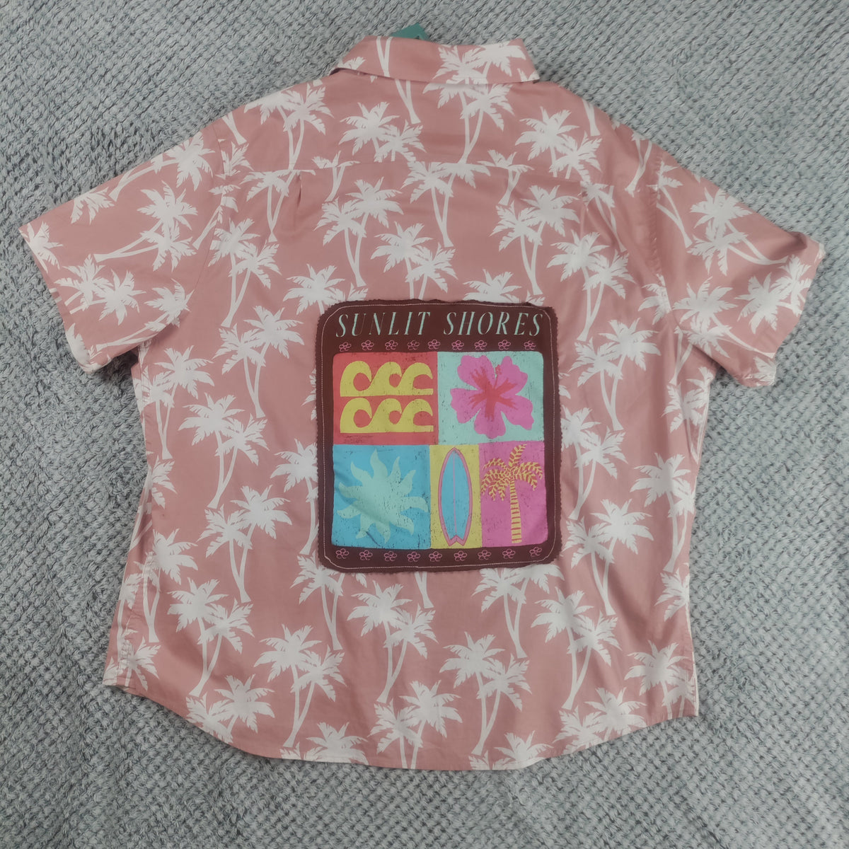 A pink short-sleeve button-up shirt with a white palm tree pattern, featuring a colorful 'SUNLIT SHORES' patch on the back, laid flat on a gray textured blanket.