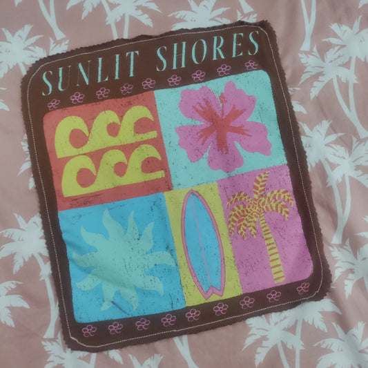 A square fabric patch with scalloped edges featuring the words 'SUNLIT SHORES' and four tropical-themed panels: yellow waves, a pink hibiscus flower, a blue palm leaf, and a surfboard with a palm tree. The patch is laid flat on a dusty pink fabric with a white palm leaf pattern.