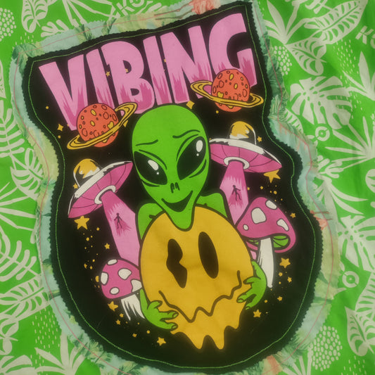 A close-up of a green patterned fabric featuring a colorful graphic patch with the word 'VIBING' in pink, a green alien holding a yellow moon, pink mushrooms, and small planets on a black background.