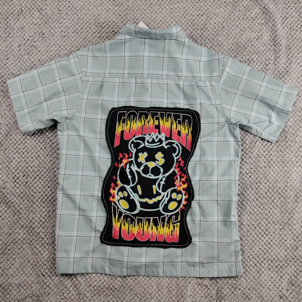 A light gray and white plaid short-sleeve button-up shirt laid flat on a textured gray blanket, featuring a large 'Forever Young' teddy bear patch on the back.