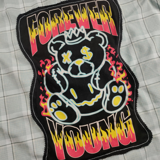 A close-up of a black embroidered patch featuring a cartoon teddy bear with yellow paw pads and a crown, surrounded by red and yellow flame-like accents, with the words 'FOREVER YOUNG' in a dripping red, yellow, and black font. The patch is laid flat on a light gray plaid fabric background.
