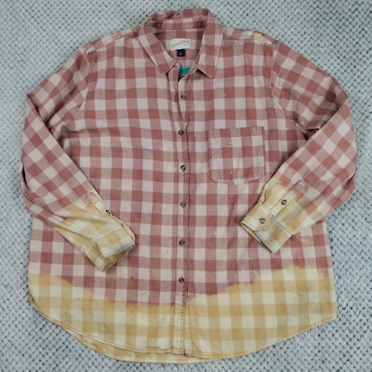 A long-sleeve button-up flannel shirt laid flat on a textured gray background. The shirt features a dusty pink and white gingham pattern on the upper body and sleeves, with a yellow and white gingham pattern on the lower hem and turned-back cuffs.