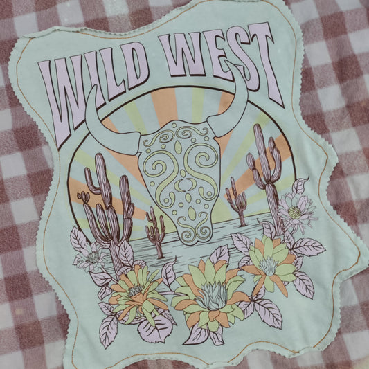 A close-up of a fabric patch with a scalloped edge featuring a 'Wild West' design. The patch includes a decorative skull, cacti, flowers, and a sunburst in muted pastel colors, laid flat on a brown and white checkered fabric background.