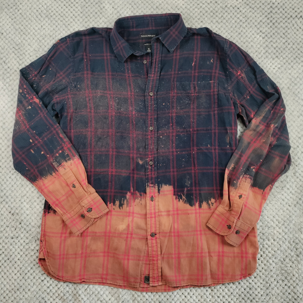A long-sleeve button-up shirt laid flat on a textured gray blanket. The shirt features a dark navy blue and red plaid pattern on the upper body, fading into a solid coral-orange color on the lower half and sleeves, with a distressed splatter effect.