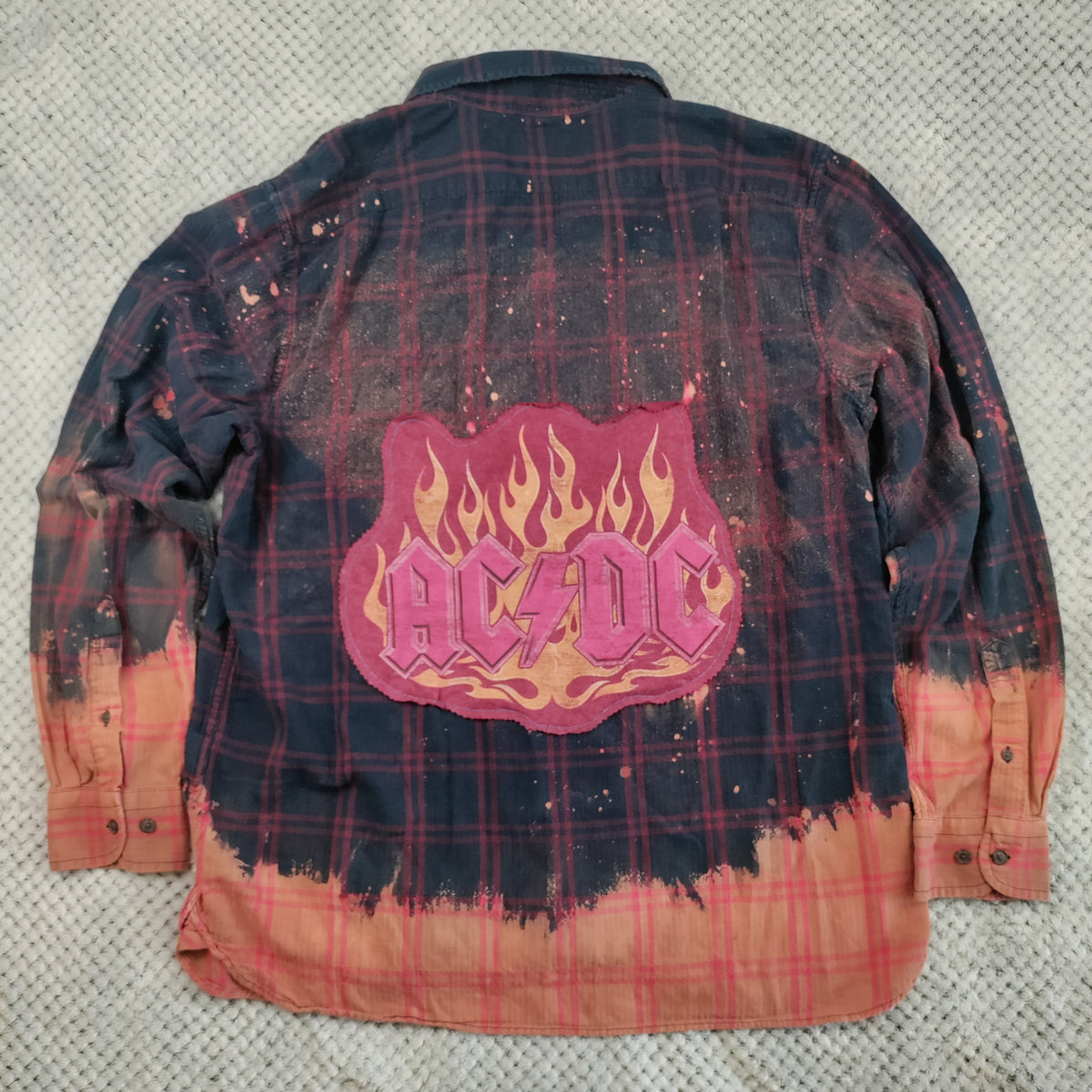 A long-sleeve AC/DC band flannel shirt laid flat on a textured gray fabric background. The back of the shirt features a dark navy and red plaid pattern with a central pink and orange flame-shaped AC/DC logo patch, and a distressed orange-brown tie-dye effect along the bottom hem and cuffs.