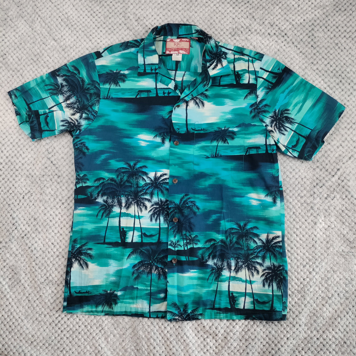 A short-sleeve button-up Hawaiian shirt with a tropical print of teal water, white sun glows, and dark palm trees, laid flat on a textured gray blanket.