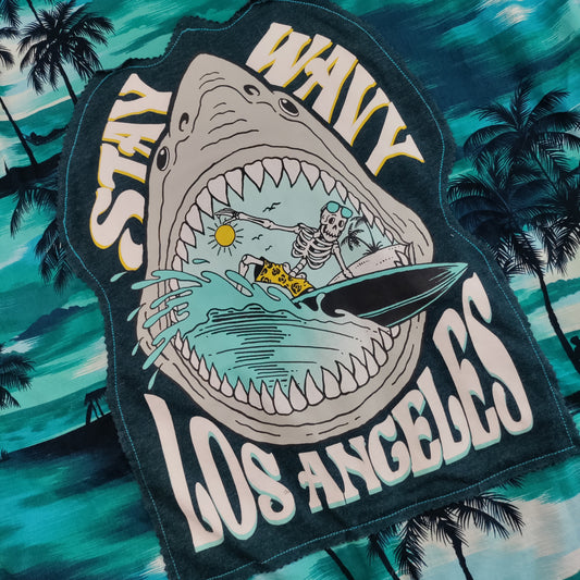 A close-up of a graphic patch featuring a shark with its mouth wide open, containing a skeleton surfing, with the text 'STAY WAVY LOS ANGELES' in white. The patch is displayed against a teal and green tropical beach background with palm trees.