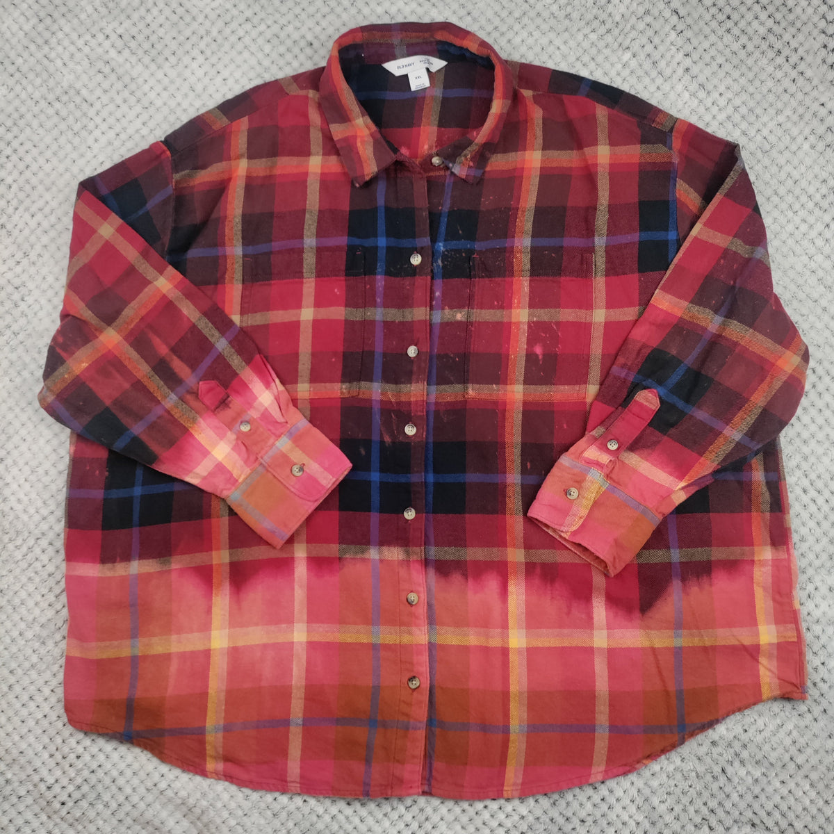A long-sleeve plaid button-up shirt in red, orange, and black, laid flat on a textured gray fabric background.
