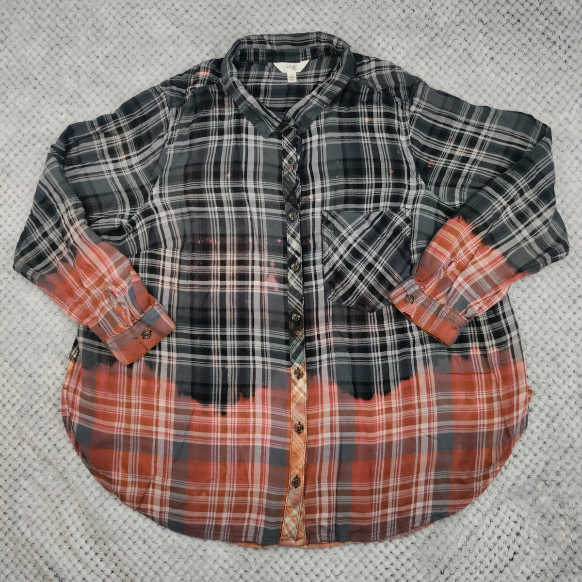 A long-sleeve plaid button-up shirt laid flat on a textured gray knit blanket. The upper portion has a dark gray, black, and white plaid pattern, while the lower portion and the turned-up cuffs feature a rust-orange and gray plaid pattern.