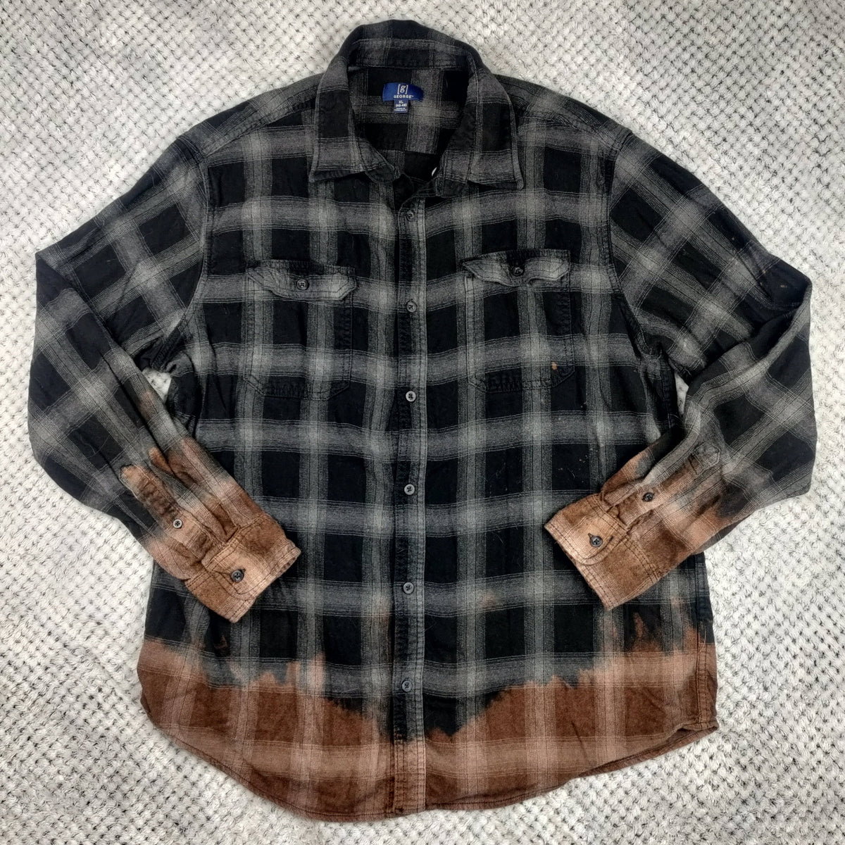 A long-sleeve plaid button-up shirt laid flat on a textured gray blanket. The shirt features a black and gray plaid pattern with a brown tie-dye or bleach-dyed effect concentrated along the bottom hem and on the turned-back cuffs.