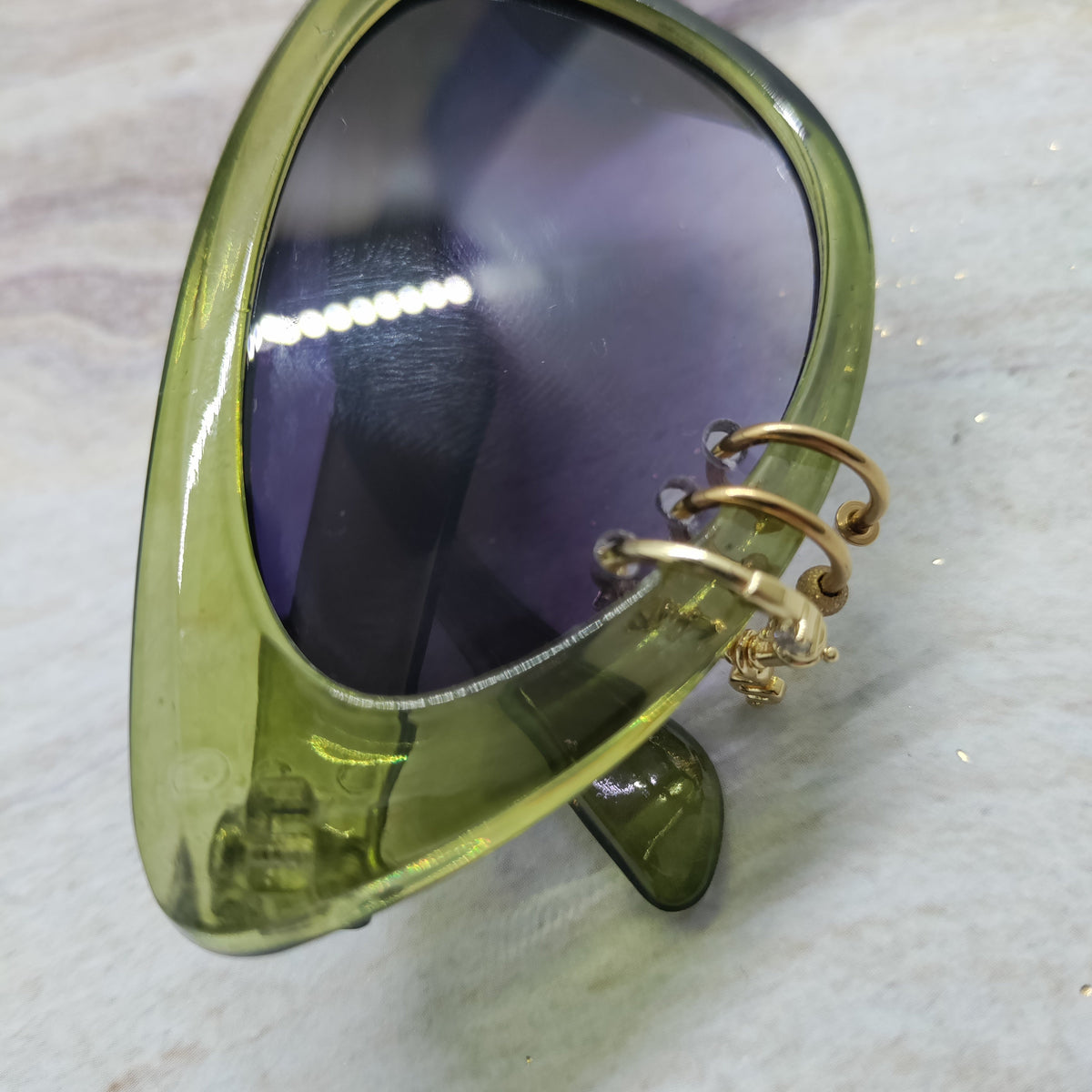 Close-up of a translucent olive green cat-eye sunglasses frame with dark tinted lenses and a gold-tone spiral ear cuff attached on the outer side, resting on a light marble-like surface.