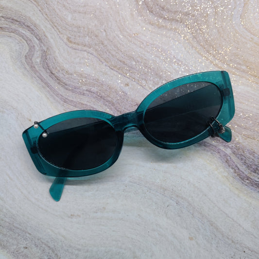 Teal translucent cat-eye sunglasses with dark lenses, displayed on a glittery white and beige marble surface.