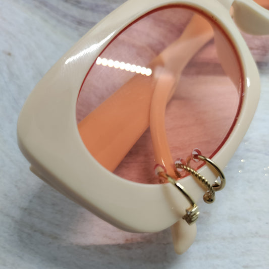 Close-up of a pair of oversized square sunglasses with a thick cream-colored frame, peach-tinted lenses, and gold hoop earrings hanging from the temple.