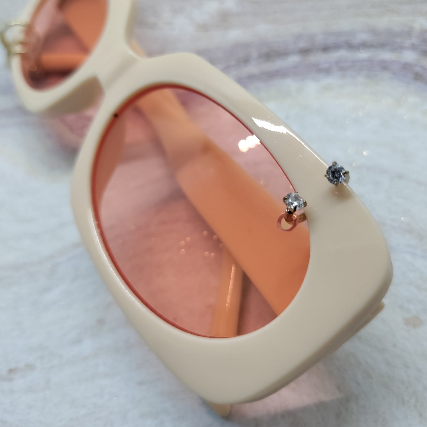 A pair of chunky square sunglasses with a cream-colored frame and peach-tinted lenses, decorated with two small silver rhinestone studs on the front right frame, resting on a light marble surface.