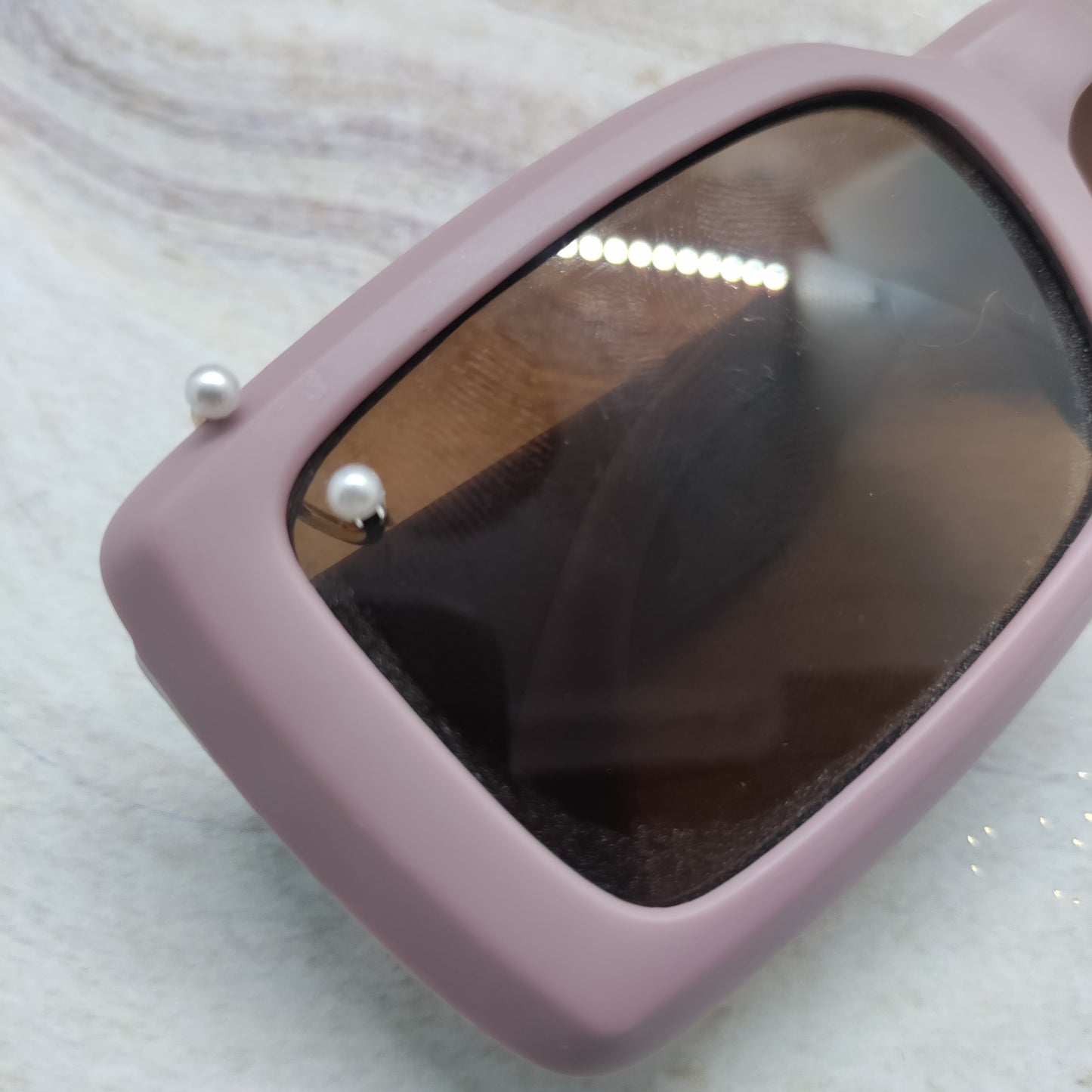A close-up of a pair of sunglasses with a dusty pink frame and dark brown tinted lenses, featuring two small white pearl-like accents on the side, resting on a light marble surface.