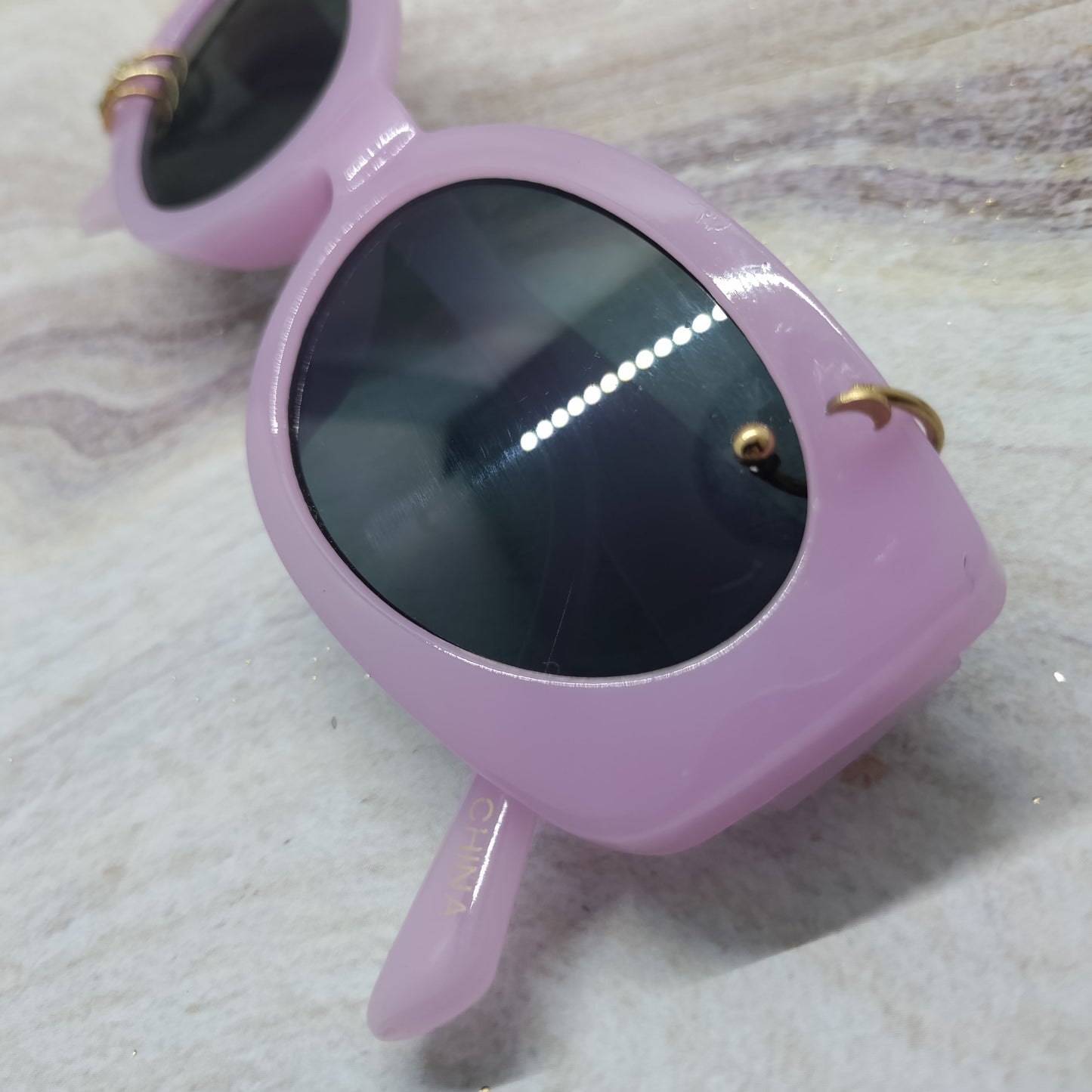 A pair of chunky, retro-style sunglasses with a glossy light pink frame and dark lenses, resting on a light-colored marble surface.
