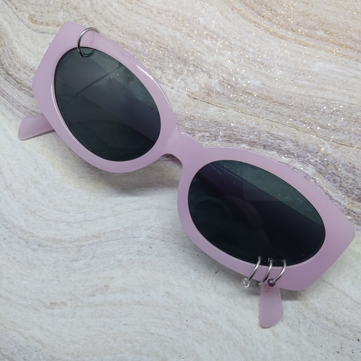 Light purple cat-eye sunglasses with dark lenses and silver hoop earrings, resting on a textured beige marble surface.