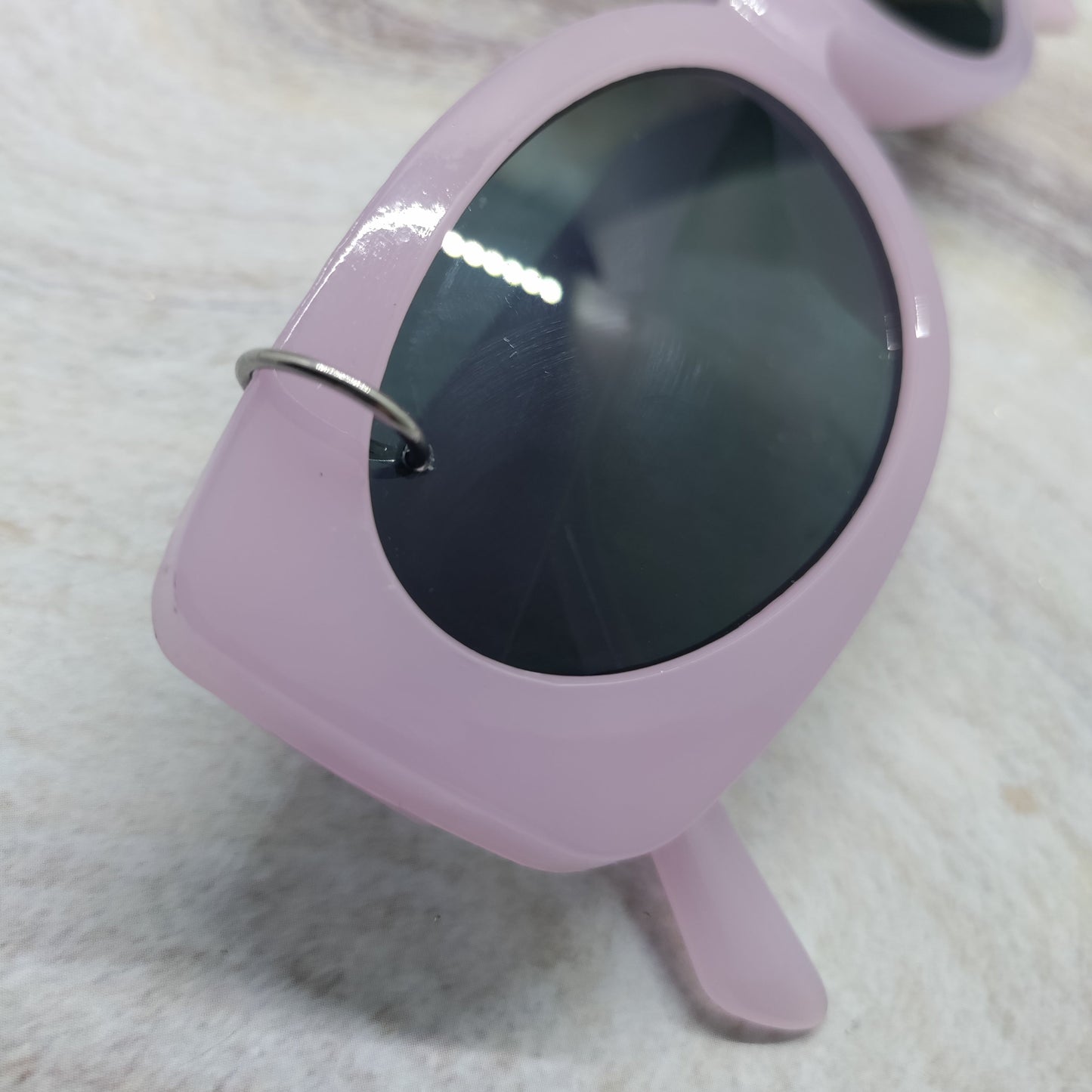 Light pink oversized sunglasses with dark tinted lenses and a small silver ring detail on the side, resting on a textured white surface.