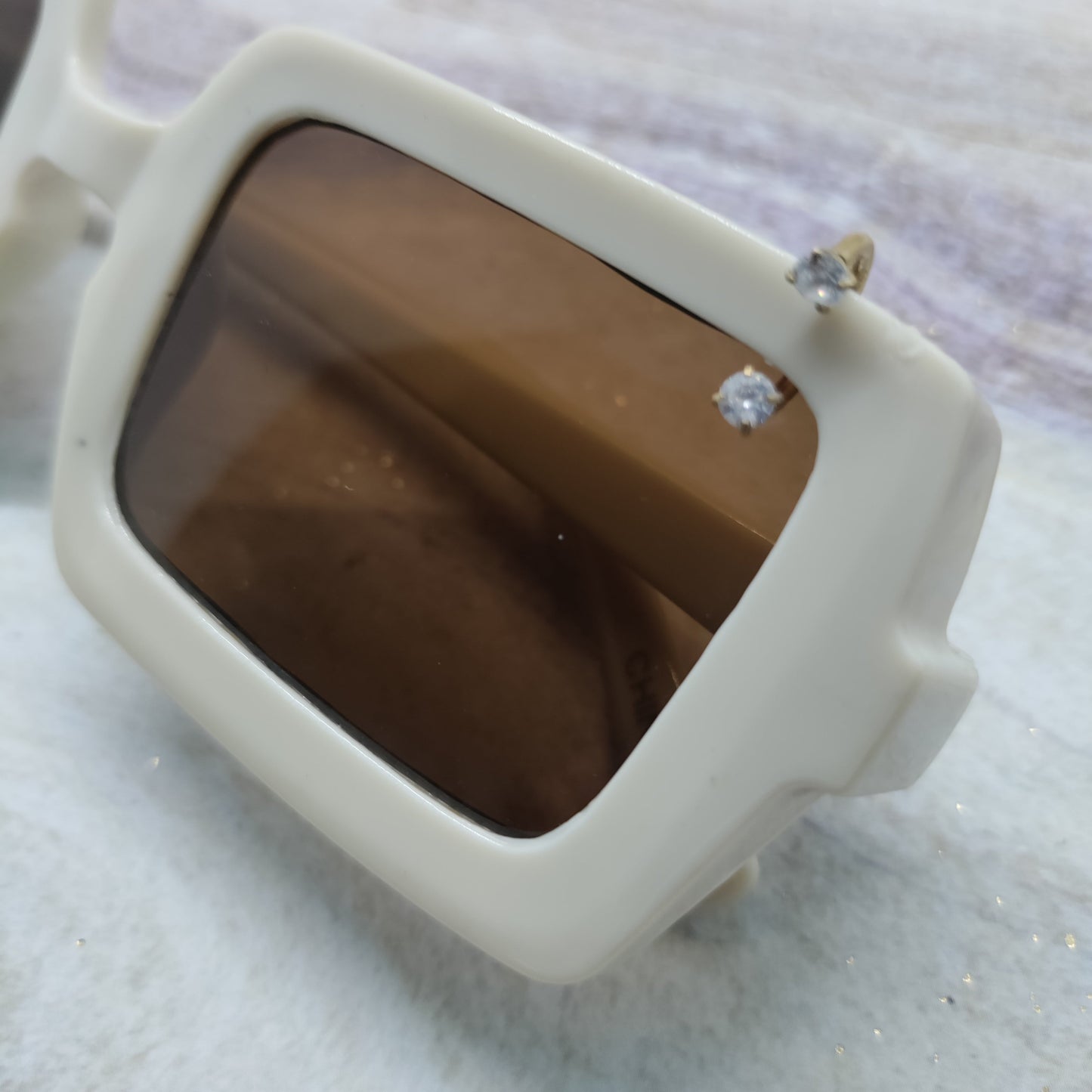 Close-up of a pair of oversized square sunglasses with a thick off-white frame and dark brown tinted lenses, resting on a light marble-like surface.