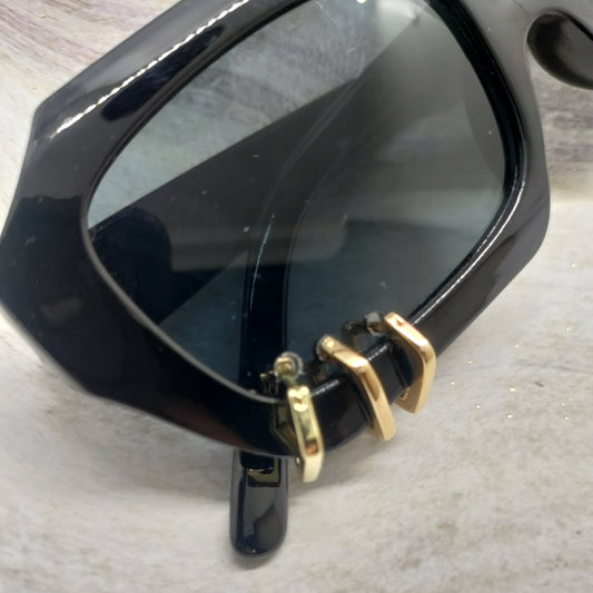 Close-up of a black acetate sunglasses frame with dark tinted lenses and gold metal hinge hardware, resting on a light textured surface.