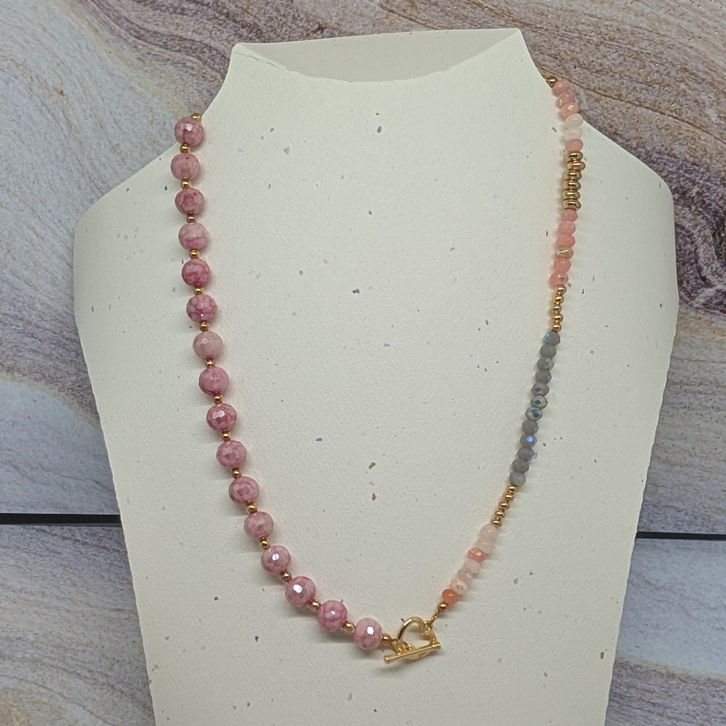 Necklace with pink beads and a heart charm on a white stand against a marble background