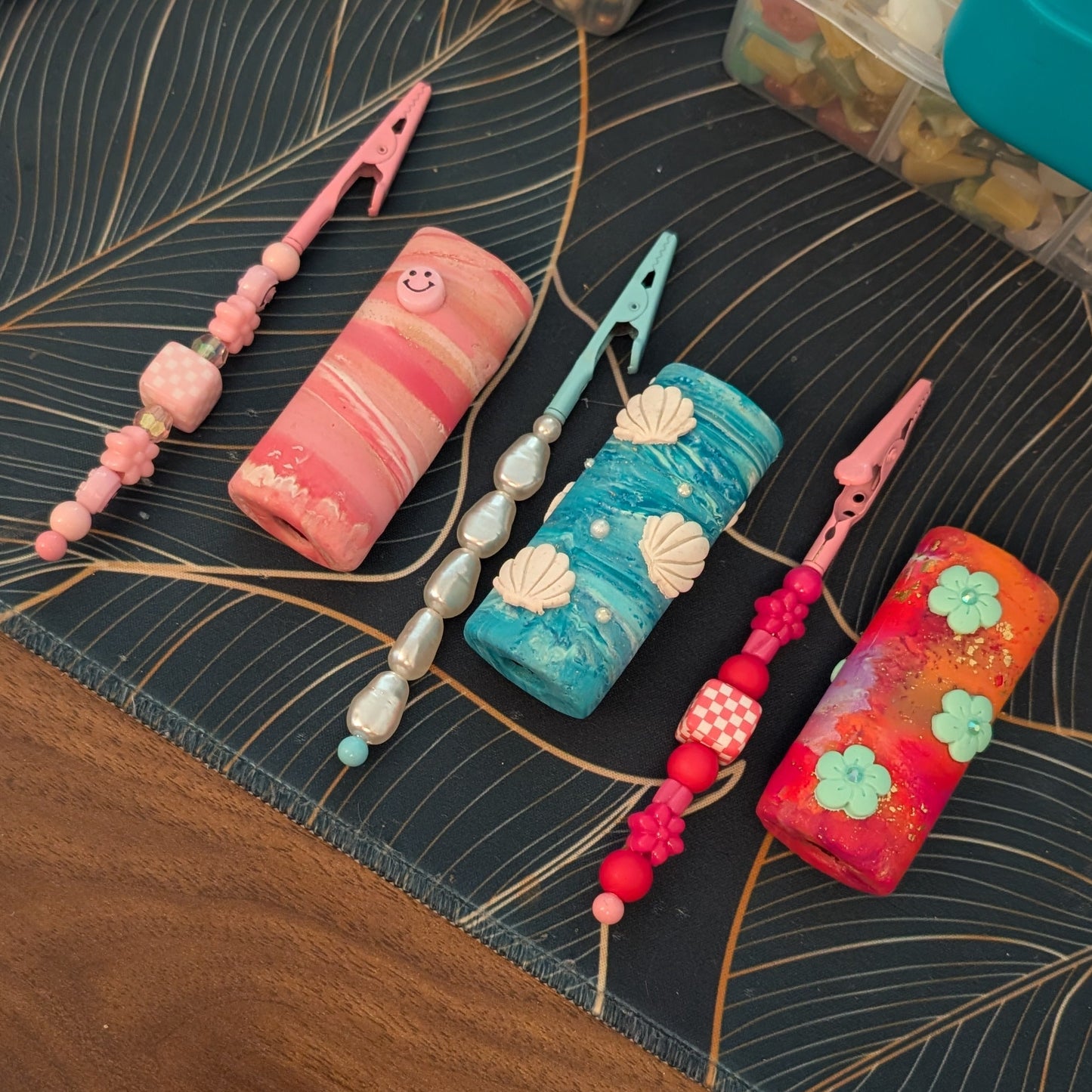 A set of colorful, decorative clay lighter case covers and matching clips, laid out on a dark patterned mat over a wooden table.