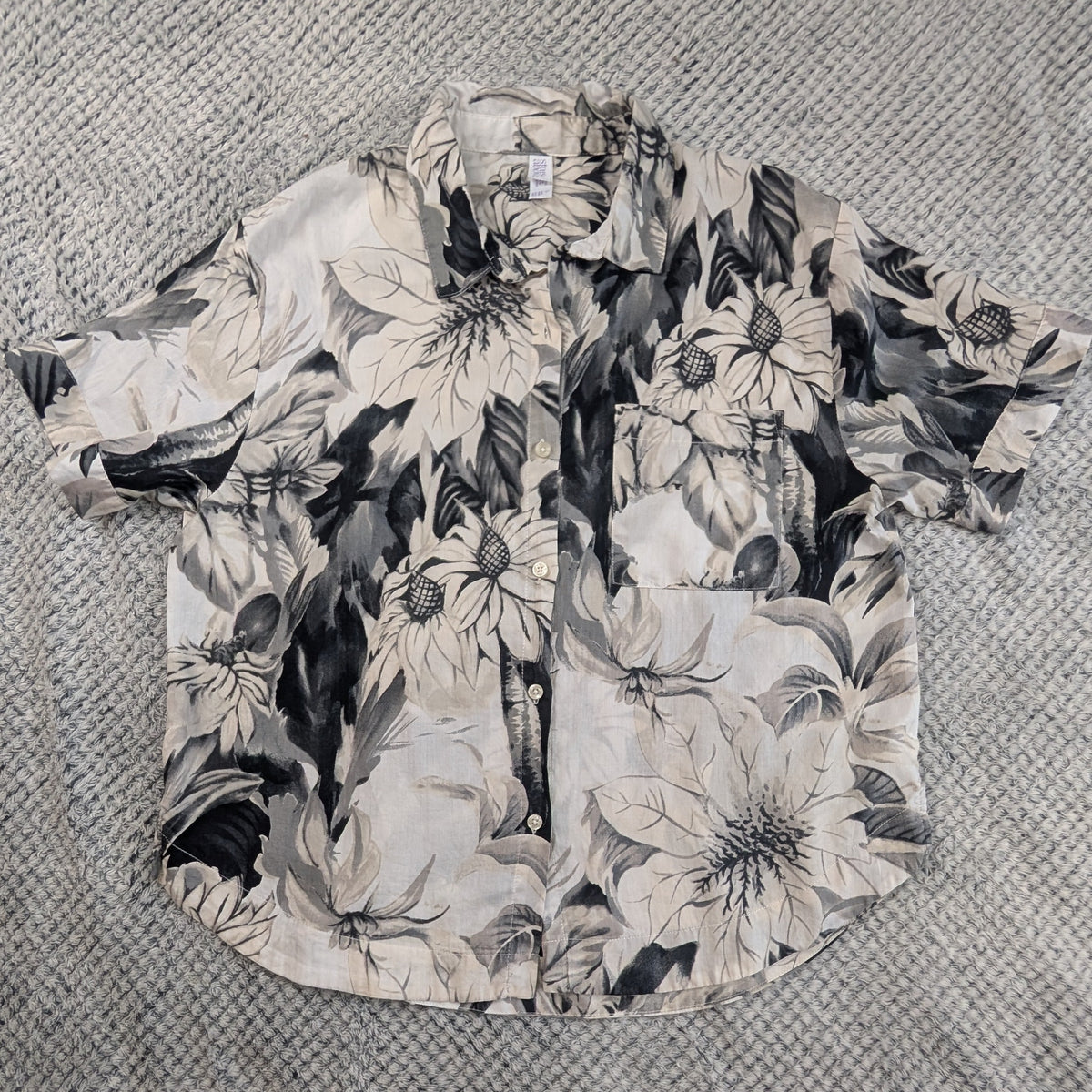 A short-sleeve button-up Hawaiian shirt with a black and white floral print, laid flat on a textured gray blanket.