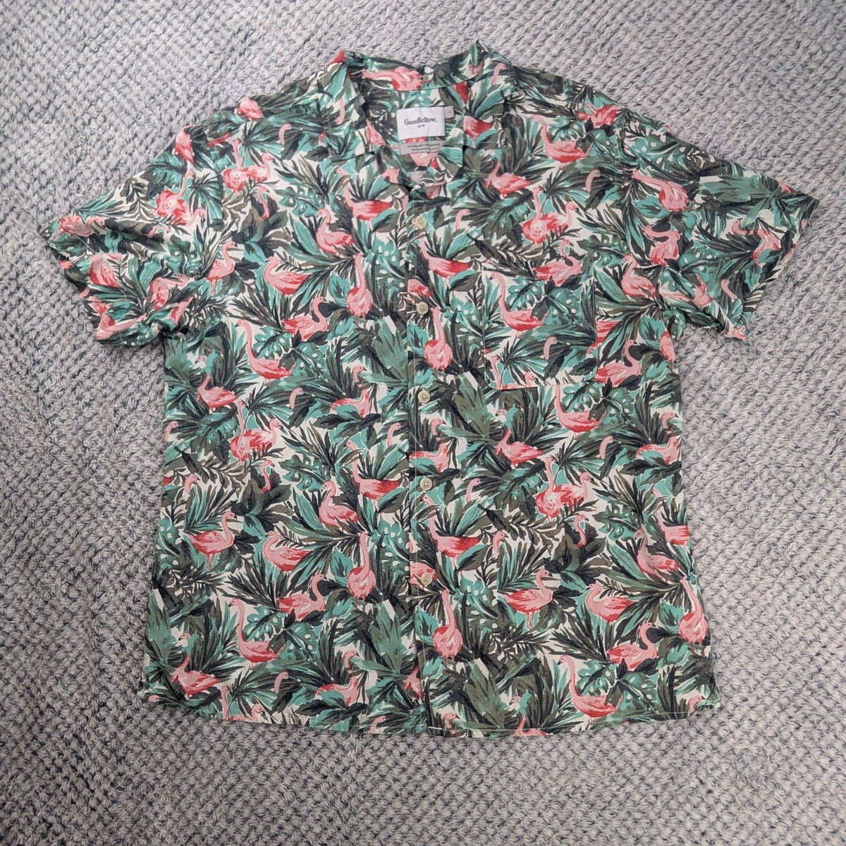 A short-sleeve button-up camp shirt with a pink flamingo and green palm leaf pattern, laid flat on a gray textured rug.