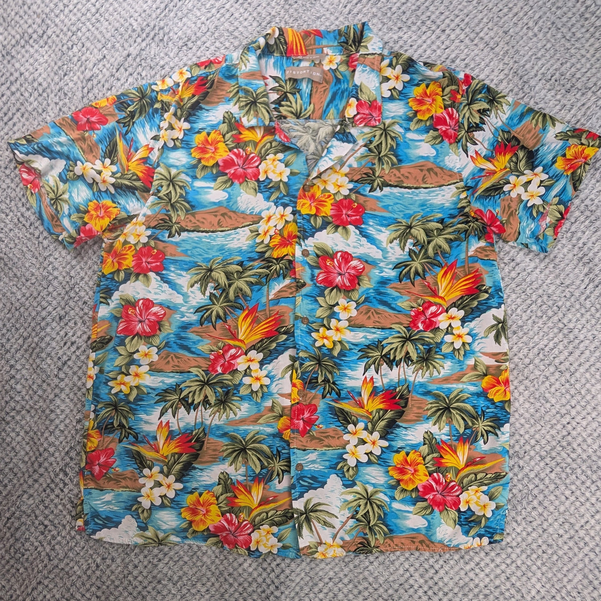 A short-sleeve Hawaiian button-up shirt with a colorful tropical print featuring palm trees, hibiscus flowers, and waves, laid flat on a gray textured blanket with bare feet visible at the bottom edge.